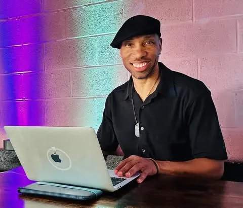 James Valentino smiling behind a laptop against a color-lit brick wall.